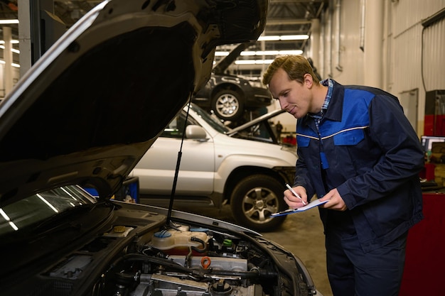 젊은 전문 정비사가 자동차 수리점에서 열린 후드의 엔진을 검사하면서 클립보드에 글을 씁니다.