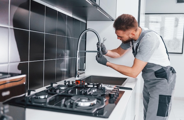 Young professional plumber in grey uniform fixing water tap on the kitchen