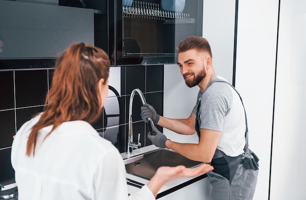 Young professional plumber in grey uniform shows results of work for housewife on the kitchen