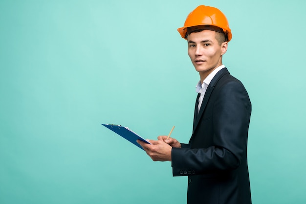 A young professional wearing a hard hat and carrying a clipboard on blue background