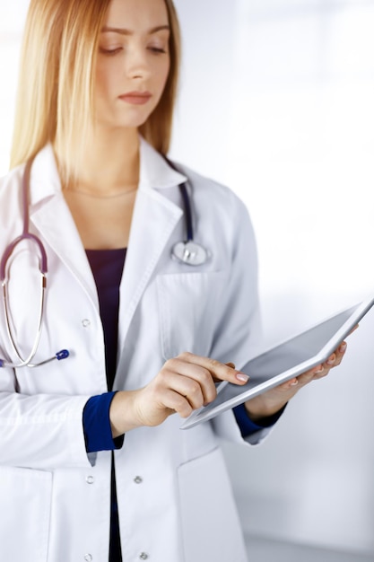 Young professional woman-doctor is using a tablet computer, while standing in a sunny clinic. Portrait of beautiful female physician at work. Perfect medical service in hospital. Medicine concept.