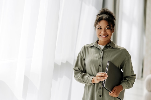 Young professional woman holding laptop smiling in office hallway