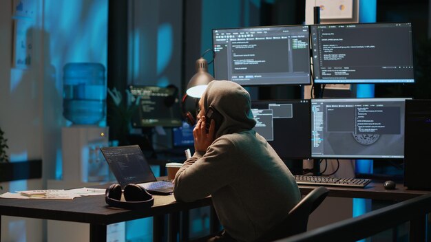 Young programmer answering phone call in office, talking on smartphone and coding user interface. Having remote conversation on mobile phone line, using html script on terminal window.