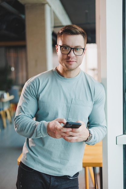 Young promising male manager in casual clothes standing by the window in the workspace and looking at the city Bright modern office in a skyscraper