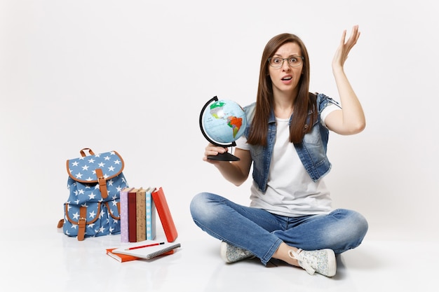 Young puzzled dissatisfied woman student in glasses holding world globe spreading hands sitting near backpack, school books isolated