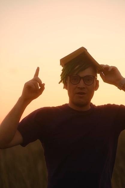 Young puzzled man in glasses with dreadlocks holding book above\
head as roof raises his index finger up invented idea in nature on\
sunset concept of roof of knowledge genius thought