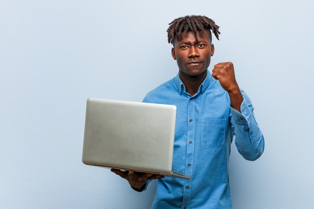 Young rasta black man holding a laptop showing fist to camera, aggressive facial expression.