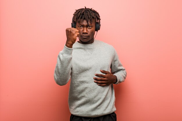 Young rasta black man listening to music with headphones showing fist to camera, aggressive facial expression.