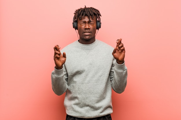 Young rasta man listening to music with headphones crossing fingers for having luck