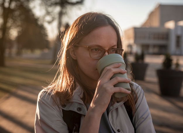 手に再利用可能なコーヒーカップ、バックライト付きのエコマグからコーヒーを飲むアイウェアの若い本物の率直な女性。