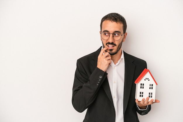 Young real estate agent man holding a model house isolated on white background relaxed thinking about something looking at a copy space.