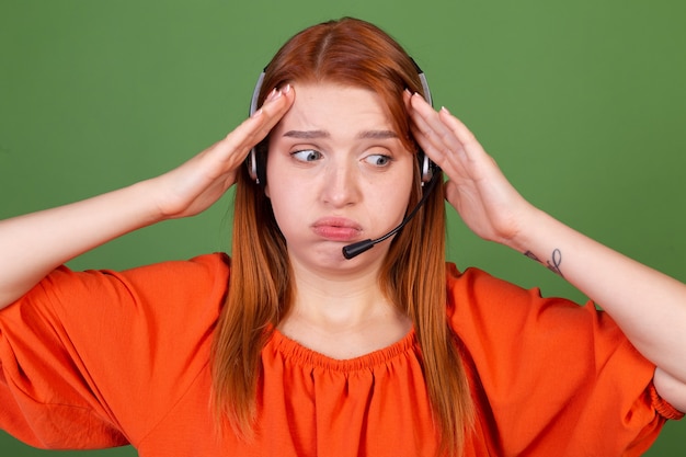 Young red hair woman in casual orange blouse on green wall manager call centre help line worker with headphones talk tired exhausted