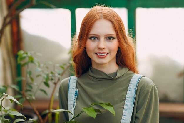 Giovane ritratto rosso della lavoratrice agricola dei capelli che osserva e che sorride in serra. concetto di giardinaggio domestico di primavera.