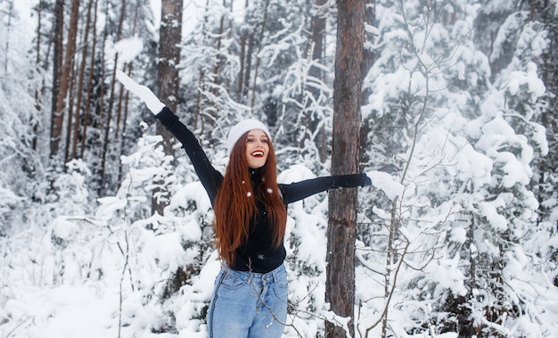 Молодая рыжеволосая девушка с длинными рыжими волосами на зимнем фоне. Рыжая женщина в белой шляпе и вязаных перчатках на фоне зимней природы.