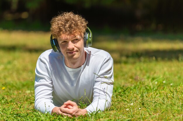 Photo young red-haired man in headphones lies on the green grass on a sunny day