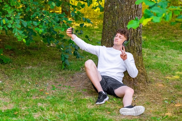 Young red-haired man in headphones sitting leaning on a tree taking a selfie