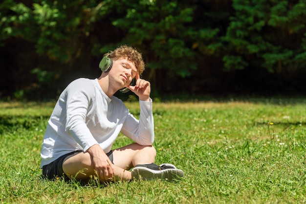 Giovane uomo dai capelli rossi con le cuffie che si siede sull'erba verde in una giornata di sole