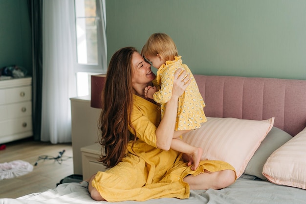 Young red-haired mother plays with her little cheerful daughter sitting on the bed