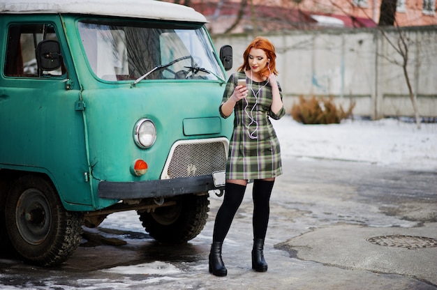 Young red haired woman lady with mobile cell phone and headphones, wearing on checked dress in old vintage minivan bus. 