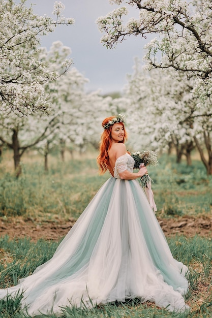 Young red-haired woman in a luxurious dress is standing in a blooming garden