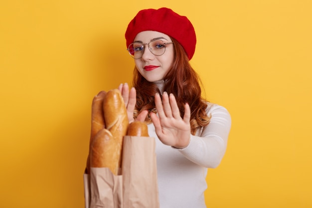 Young red haired woman with curls refuses from fresh bread baguette offering from unknown person on yellow
