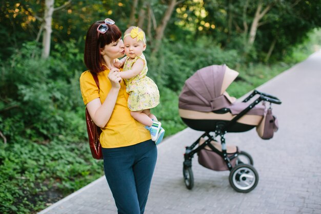 黄色のシャツを着た若い赤毛の女性が公園で夏の明るい日にベビーカーで小さな赤ちゃんと一緒に歩いています。