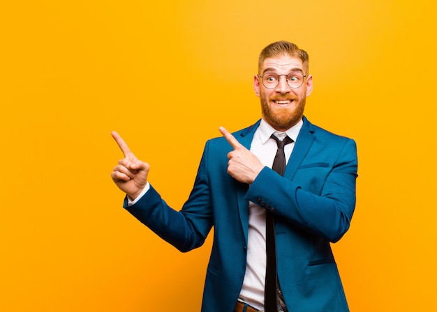 Young red head businessman feeling joyful and surprised, smiling with a shocked expression and pointing to the side