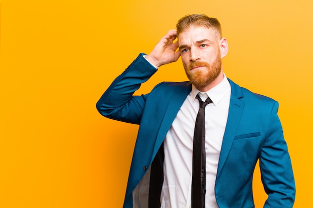 Young red head businessman feeling puzzled and confused, scratching head and looking to the side on orange