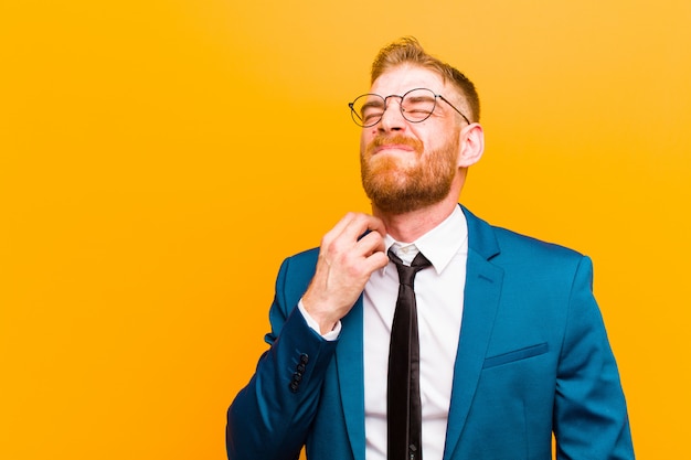 Young red head businessman feeling stressed, frustrated and tired, rubbing painful neck, with a worried, troubled look against orange 