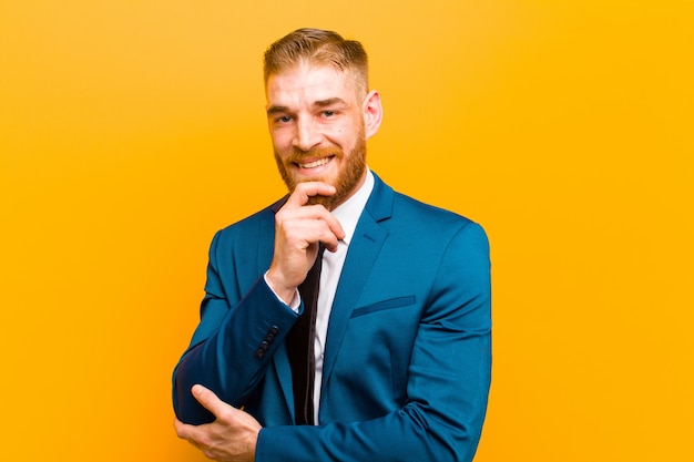 Young red head businessman looking happy and smiling with hand on chin, wondering or asking a question, comparing options