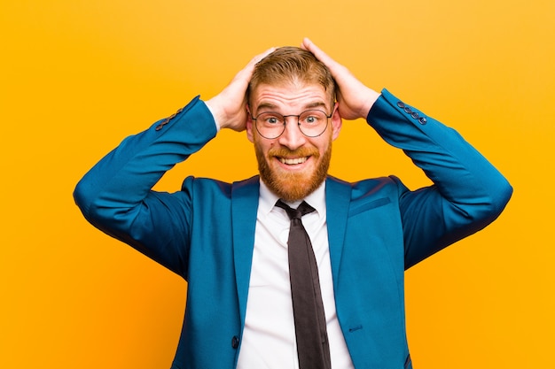 Photo young red head businessman raising hands to head, open-mouthed, feeling extremely lucky, surprised, excited and happy on orange