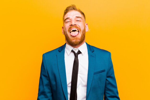 Young red head businessman with cheerful, carefree, rebellious attitude, joking and sticking tongue out, having fun against orange 