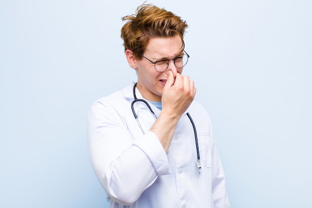 Young red head doctor feeling disgusted, holding nose to avoid smelling a foul