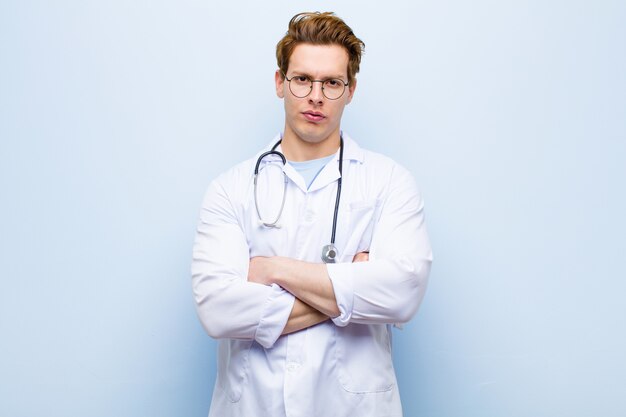 不満と失望、深刻な、イライラ、怒っている青い壁に組んだ腕に見える若い赤ヘッド医師