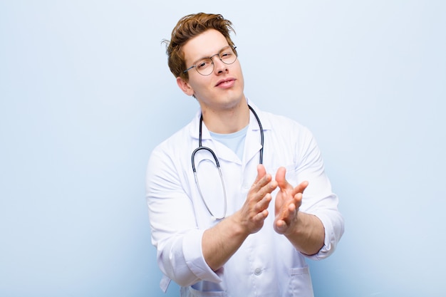 Young red head doctor feeling happy and successful, smiling and clapping hands, saying congratulations with an applause against blue wall