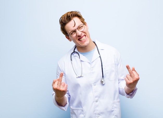 Young red head doctor feeling provocative, aggressive and obscene, flipping the middle finger, with a rebellious attitude against blue wall