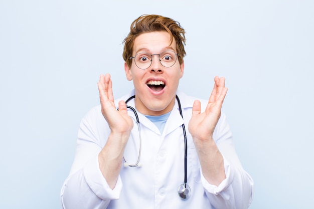 Young red head doctor feeling shocked and excited, laughing, amazed and happy because of an unexpected surprise against blue wall