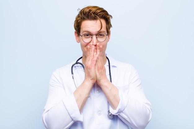 Young red head doctor happy and excited surprised and amazed covering mouth with hands giggling with a cute expression against blue wall