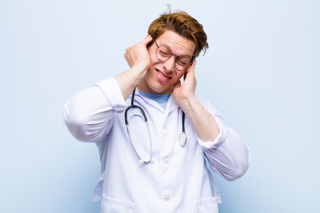 Young red head doctor looking angry, stressed and annoyed, covering both ears to a deafening noise, sound or loud music against blue wall