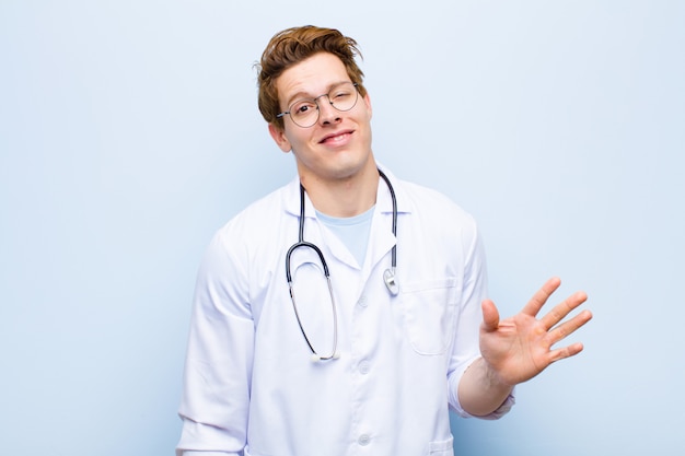 Young red head doctor smiling happily and cheerfully, waving hand, welcoming and greeting you, or saying goodbye  blue wall