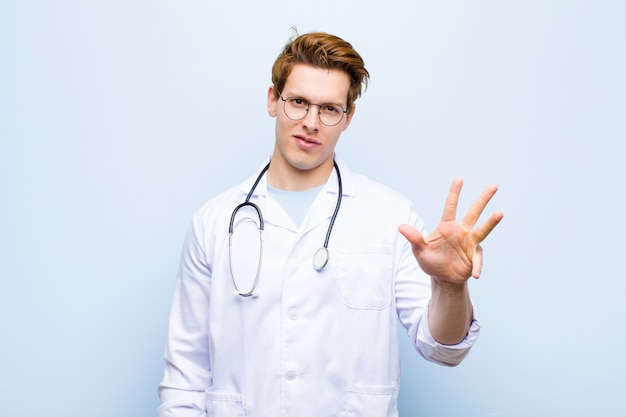 Young red head doctor smiling and looking friendly, showing number four or fourth with hand forward, counting down against blue wall