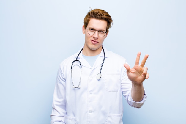 Young red head doctor smiling and looking friendly, showing number three or third with hand forward, counting down against blue wall