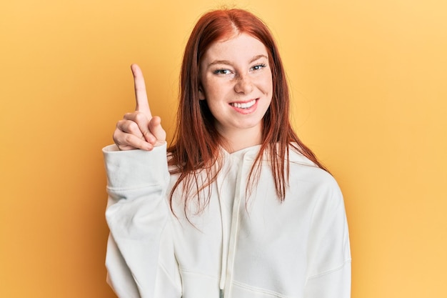 カジュアルなスウェットシャツを着た若い赤い頭の女の子が自信と幸せで笑顔を浮かべて指を指して上を指しています