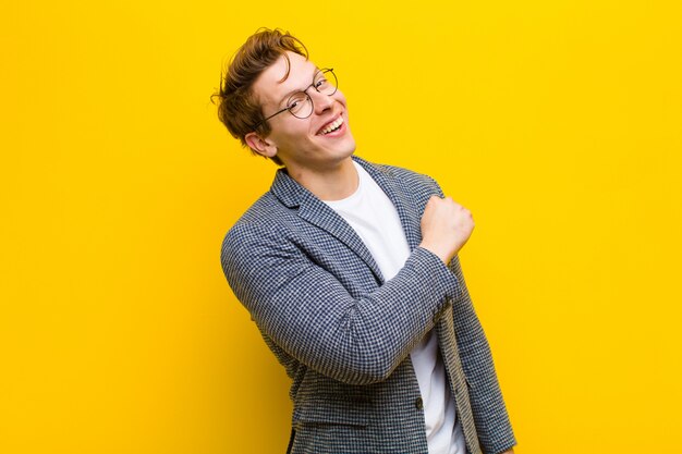 Young red head man feeling happy, positive and successful, motivated when facing a challenge or celebrating good results against orange background