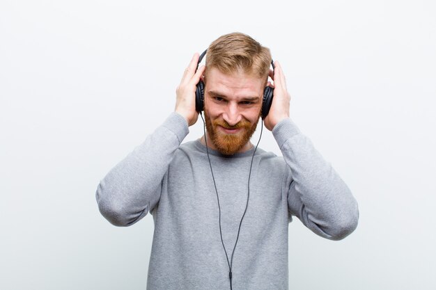 Young red head man listening music with headphones against white 