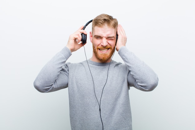 Young red head man listening music with headphones   white wall