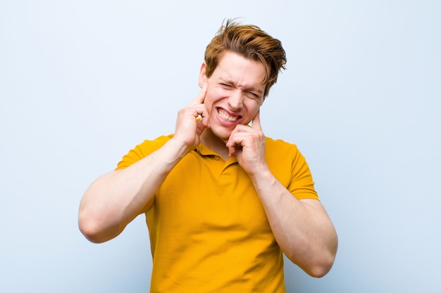 Young red head man looking angry, stressed and annoyed, covering both ears to a deafening noise, sound or loud music on blue wall