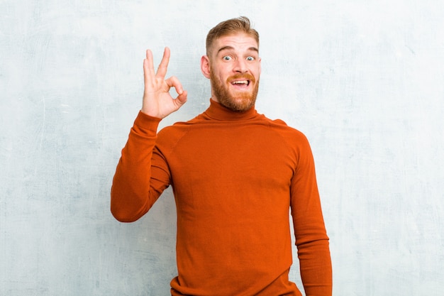 Young red head man wearing turtle neck feeling successful and satisfied, smiling with mouth wide open, making okay sign with hand against cement wall