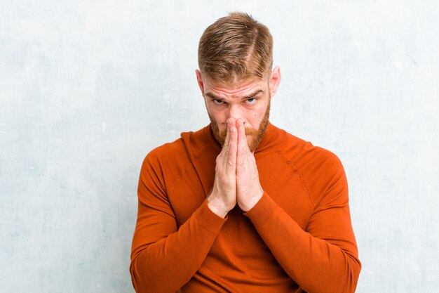 Young red head man wearing turtle neck feeling worried, hopeful and religious, praying faithfully with palms pressed, begging forgiveness against concrete wall