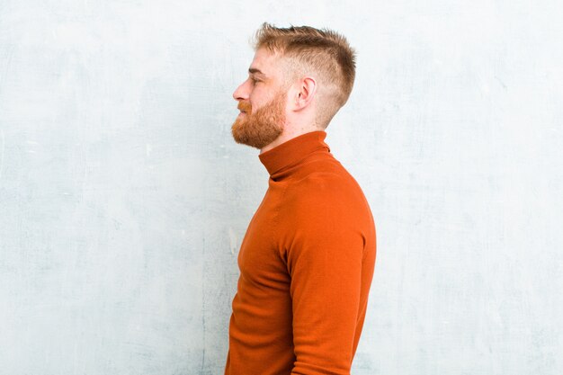 Photo young red head man wearing turtle neck on profile view looking to copy space ahead, thinking, imagining or daydreaming against concrete wall